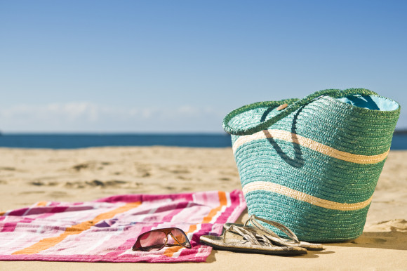 A straw beach bag placed next to a towel, a pair of sunglasses and some flip flops with the ocean in the distance