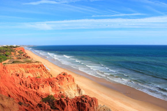 A high up view of the picturesque coastline of Praia da Falésia Beach in Portugal with red cliffs in the background and rippling blue waters