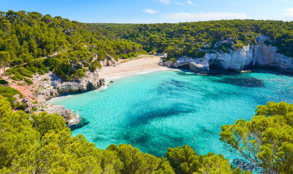 A view of the striking Mitjaneta Beach on the island of Menorca in Spain with shimmering turquoise waters and greenery