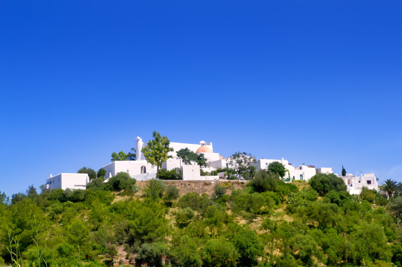 Whitewashed church on a hill in Santa Eulalia, Ibiza with a blue sky as the backdrop and green trees in the surroundings