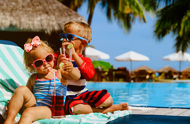 Two young children in sunglasses sitting on a sun loungers and drinking beverages next to a swimming pool