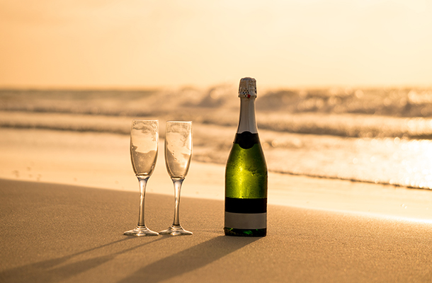 A unopened bottle of champagne on a beach with two champagne flutes