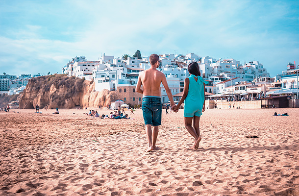 Couple walking away from the camera on a beach in Albufeira, Portugal. The whitewashed buildings of Albufeira town form the background.